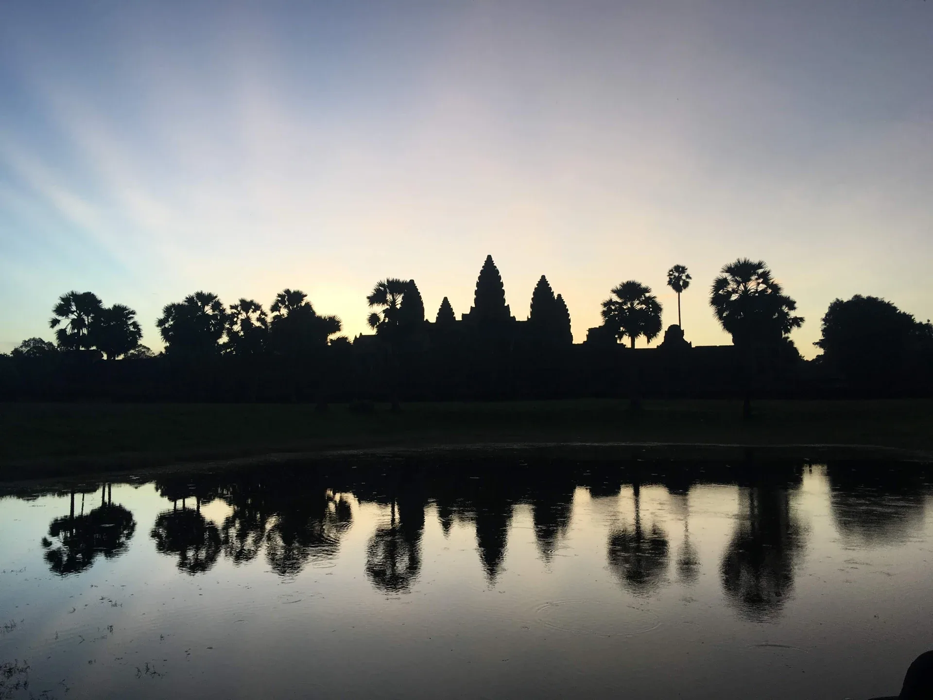 As torres de Angkor Wat e as palmeiras se refletem em um lago ao amanhecer.