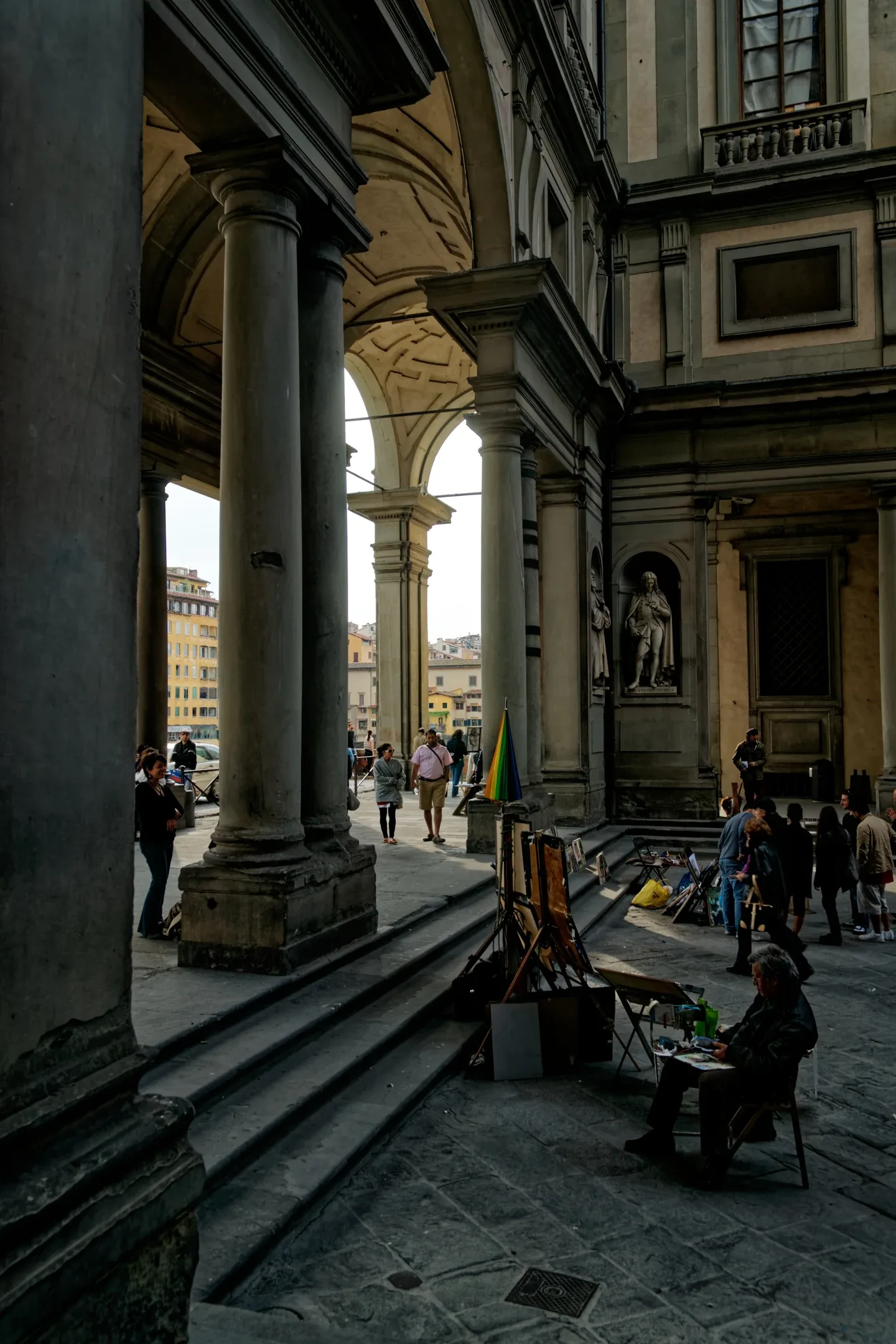 Søjler og arkader i Uffiziernes portiko med forbipasserende og kunstnere, der har slået sig ned på Arno-siden.